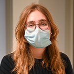 White female (Claire Manger) with long strawberry-blond hair wearing glasses, a dark-colored shirt and a surgical mask.